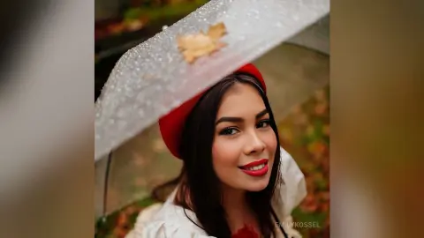 Smiling woman in a red beret holding a clear umbrella amidst autumn leaves in a cozy fall setting.