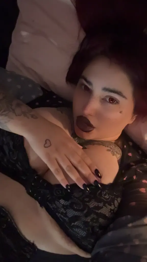 belledarkgoddess with long red hair and dark lipstick, wearing a black choker and sheer black top, looking directly at the camera.