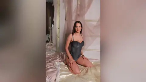 Smiling woman in a sleek grey bodysuit sitting on a luxurious satin bed in a dreamy, romantic bedroom setting.