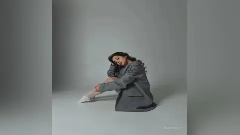 Young woman in a grey oversized blazer and white socks sitting on a grey floor with a moody, minimalist aesthetic.