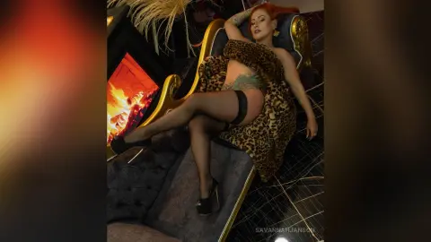 Redheaded woman in leopard print robe and black stockings lounging on a velvet sofa by a warm fireplace.