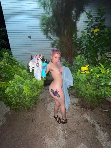 Nude woman with pink hair holding a blue water bottle in a lush garden setting at night.