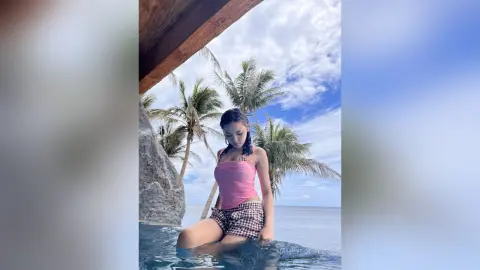 NancyLoppez sits on the edge of a swimming pool wearing a pink tank top and plaid shorts, surrounded by palm trees.