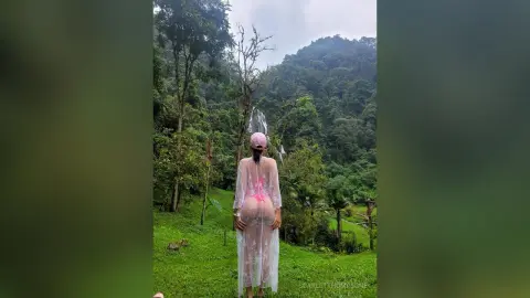 Woman in a sheer white lace cover-up standing in a lush green forest landscape.