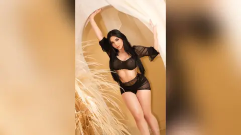 IvyStuart poses in a black sheer mesh outfit with long black hair against a light tan background with dried pampas grass.
