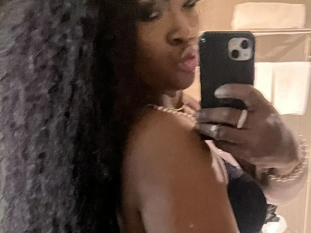 Glamorous woman in a black outfit and gold jewelry taking a moody mirror selfie in a dimly lit room.