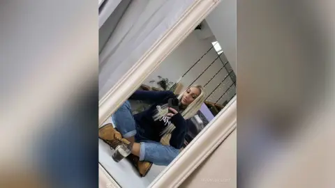 Young woman in a navy sweatshirt and blue jeans taking a mirror selfie in a bright, modern room.