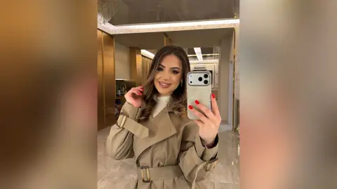 Glamorous woman in a beige trench coat taking a mirror selfie in a modern, luxury interior setting.