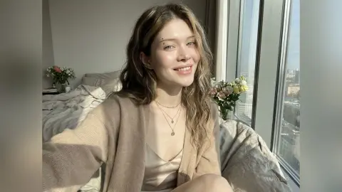 MaryLico smiles while sitting on a bed in a tan cardigan and light camisole near a window.