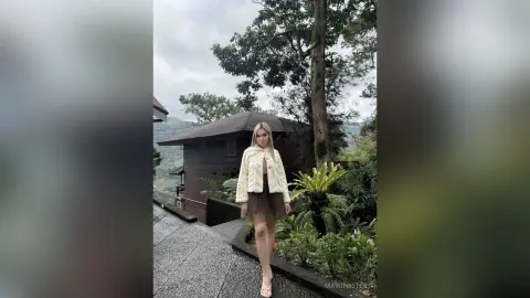 Blonde woman in a cream jacket and black skirt standing on a stone path near a rustic cabin in a lush forest.