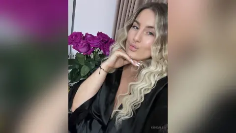 Blonde woman in a black satin robe smiling near purple roses in a soft, dreamy indoor setting.