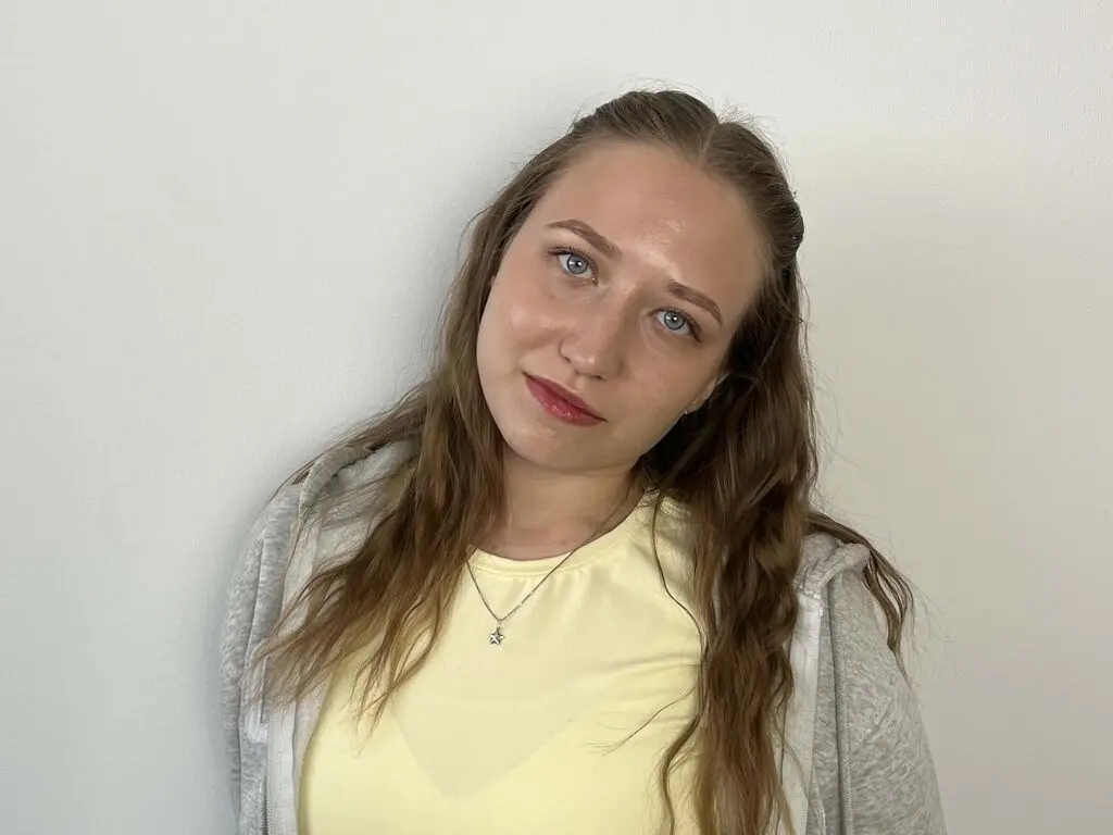 Young woman with long wavy hair wearing a yellow shirt and grey hoodie against a plain white wall.