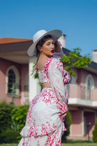 Elegant woman in a white and pink floral maxi dress and sun hat posing outdoors near a pink villa.