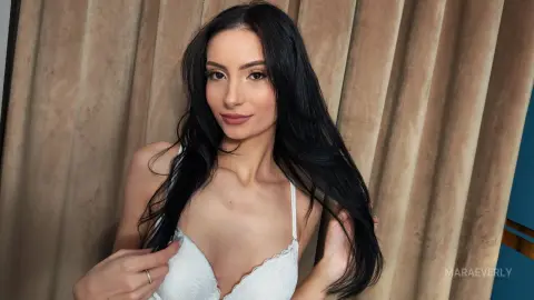 Beautiful woman with long black hair wearing a white lace bra in a soft, neutral indoor setting.
