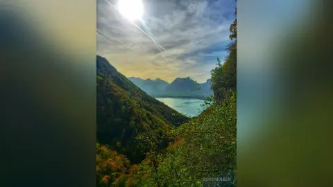 RossiNoleen overlooks a vast, sunlit valley and a blue lake surrounded by green and autumn-colored mountains.