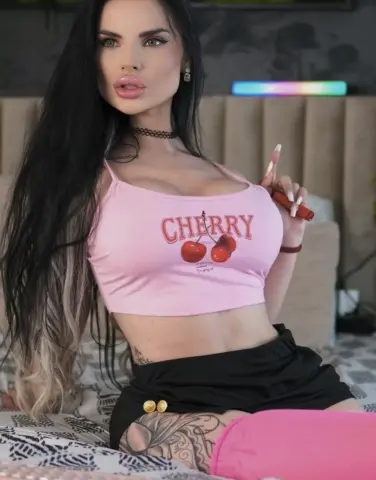 Young woman with long dark hair wearing a pink cherry crop top and black shorts sitting on a bed.