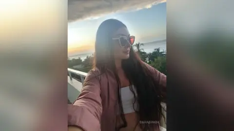 Young woman in sunglasses and pink shirt enjoying a sunset view on a tropical balcony.