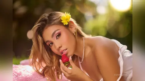 Beautiful woman with a yellow flower in her hair eating a strawberry in a sunny, dreamy outdoor setting.