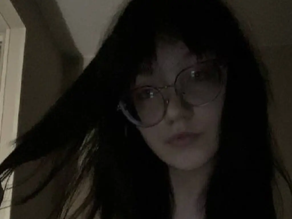 Moody portrait of a young woman with dark hair and round glasses in dim indoor lighting.
