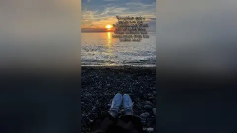 SofiMils sits on a rocky beach at sunset, wearing white sneakers and dark pants, looking out over the ocean waves.