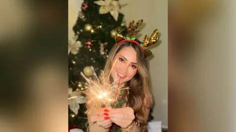 Smiling woman in reindeer headband holding a sparkling firework in front of a festive Christmas tree.