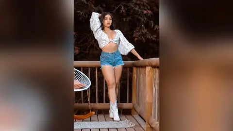 Young woman in a white floral crop top and denim shorts posing confidently on a wooden balcony.