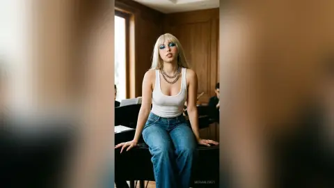 Blonde woman in white tank top and blue jeans sitting on a piano in a moody, sunlit room.