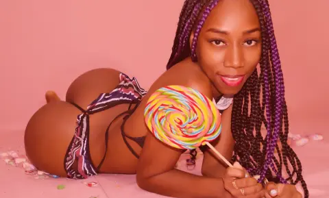 Smiling Black woman with purple braids in patterned bikini holding a colorful swirl lollipop against pink background.