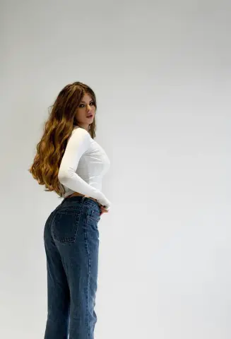 Young woman with long wavy hair wearing a white long-sleeve top and blue jeans against a white background.