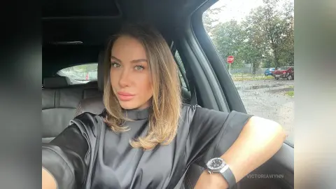 VictoriaWynn poses in a car wearing a black satin top, looking directly at the camera with her long brown hair down.