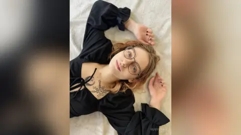 Young woman with glasses and tattoos wearing a black long-sleeve top lying on a white bed in a cozy mood.