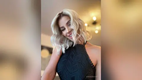 Smiling blonde woman in a black lace halter top posing in a warm, brightly lit indoor setting.