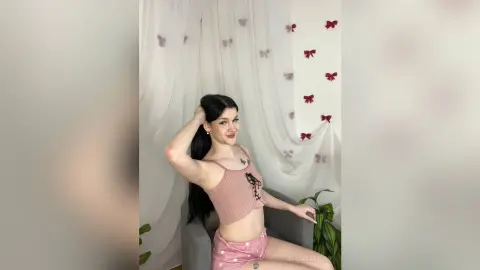 Smiling young woman in a pink ribbed crop top and shorts posing in a bright room with white curtains.