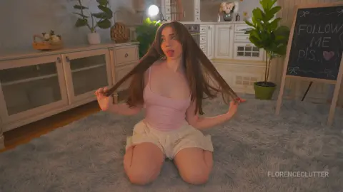 Young woman in a pink tank top and lace shorts playing with her hair on a grey rug in a cozy living room.