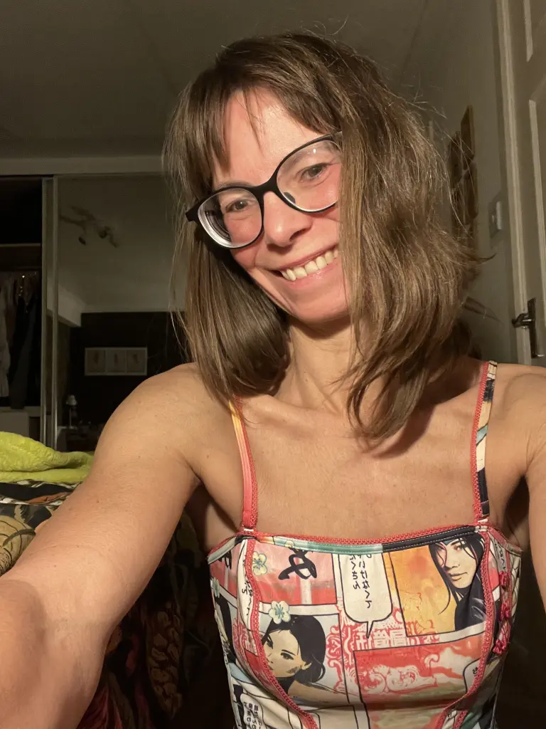Dixie_Milf smiling head-on with her brown hair styled with bangs and sunglasses resting on top of her head against a plain grey background.