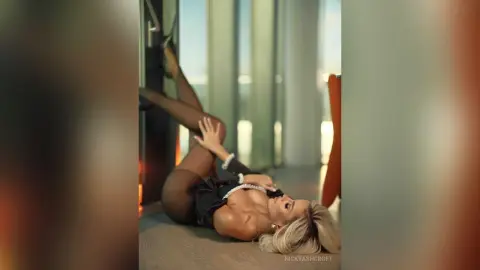Blonde woman in a black bodysuit relaxing on a floor near large windows in a bright, modern room.