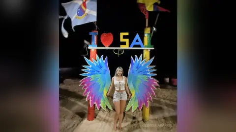 Young woman in white tank top and denim shorts posing with colorful angel wings at a night beach festival.