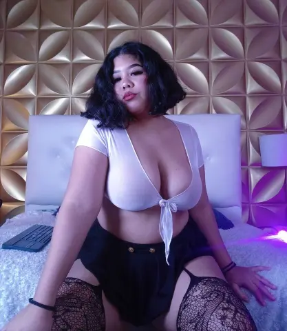 Curvy woman in a white tie-front crop top and black shorts sitting on a bed against a gold textured wall.