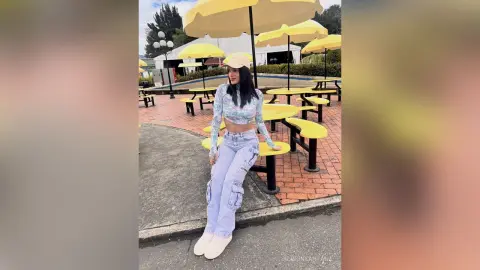 Young woman in a floral crop top, baggy jeans, and cap relaxing at an outdoor cafe with yellow umbrellas.