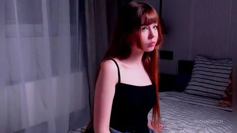 Young woman in a black tank top sitting on a bed in a dimly lit bedroom with a thoughtful expression.