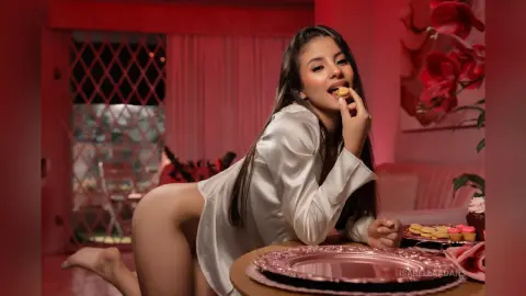 Young woman in a white satin shirt eating a snack in a cozy, warm-lit bedroom setting.