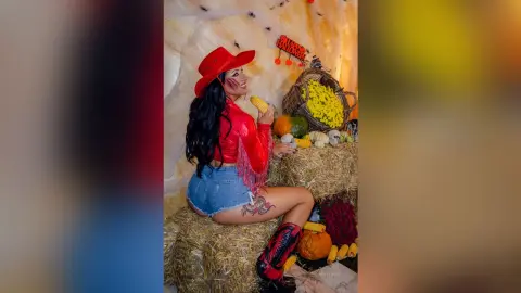 MissyJolie wears a red cowboy hat and red top while sitting on hay next to corn and pumpkins.