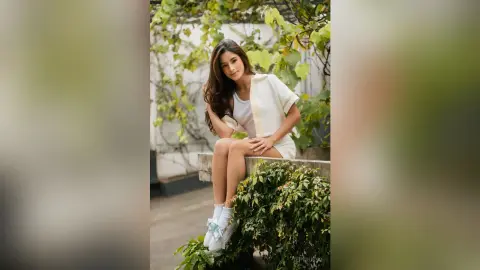 TefyRusso sits outdoors with long brown hair, wearing a white tank top and white shorts, posing near green leaves and a stone ledge.