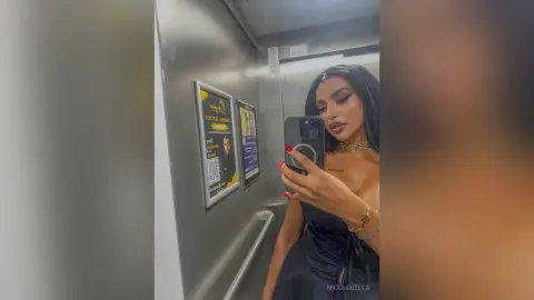 NicoleBella takes a mirror selfie in a lift, wearing a black strapless top and gold jewelry.