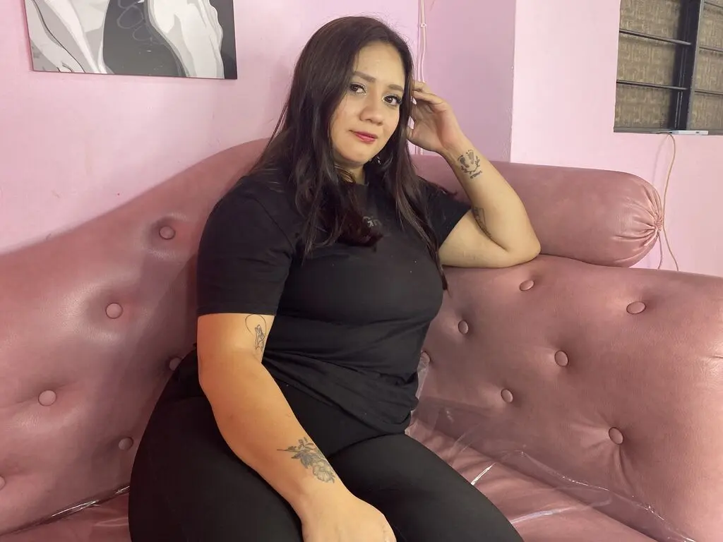 SammieRouse sits on a pink tufted couch wearing a black t-shirt and black leggings, running her hand through her long dark hair.