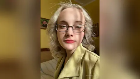 Blonde woman with glasses wearing a yellow blazer in a warm, sunlit indoor setting.