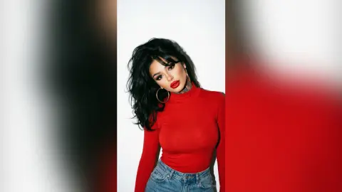 Glamorous woman with dark hair and red lipstick wearing a red turtleneck in a minimalist studio setting.