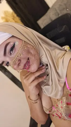 Smiling woman in gold bridal headpiece and beige hijab wearing a pink floral lace outfit.