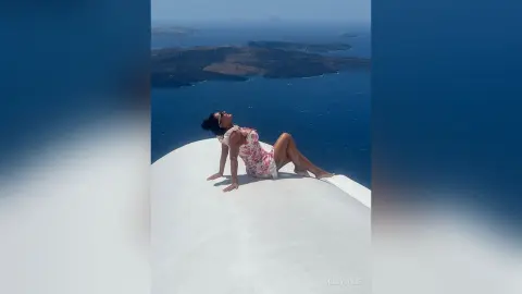 MissyJolie reclines on a white ledge overlooking a blue ocean, wearing a white floral sundress.