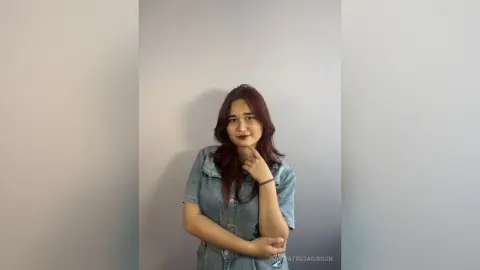 PatriciaGibson poses with her hand under her chin, wearing a blue denim shirt against a plain grey wall.